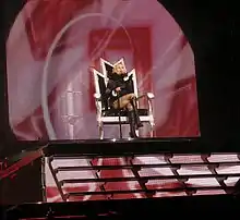 A faraway image of a blond woman sitting on a throne, atop a flight of stairs, on a stage. Two male dancers in black coat and hat, stand in front of her. A red colored backdrop is visible behind the throne.