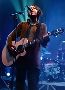 Nicholls is shown singing into a microphone while strumming a guitar.