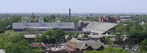 An elevated view of several buildings and the trees surrounding them. A red brick building with a sloped roof is in the foreground, and a large white football stadium is just behind it, taking up much of the center of the picture. Beyond the stadium, there is a red brick smokestack near the center of the picture, the red brick Tech Tower building on the left side bearing white letters that spell "TECH", and the red brick physics building on the right side. In the background there is a white domed building. All around these buildings are green-leafed oak trees. An overcast, light blue sky takes up the top third of the picture.