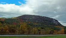 A relatively flat-topped mountain rising in the background. The trees near its base have some leaves in fall color. There is a roadway at the bottom, in the foreground.