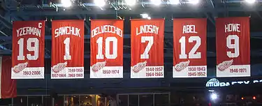 A row of six red banners. The banners, from left to right, read "Yzerman 19" "Sawchuk 1" "Delvecchio 10" "Lindsay 7" "Abel 12" "Howe 9". The Yzerman banner has a small "C" at the top right corner.