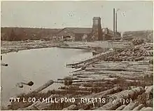 Sepia-tone photo of a pond surrounded by large logs. At the far end of the pond is a large building with a square tower and two smokestacks. Label is "T&T L CO MILL POND RICKETTS PA 1903" (i.e. Trexler and Turrell Lumber Company Mill Pond&nbsp;...)
