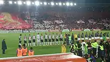 Two football teams and a refereeing team lining up in a football stadium pitch. The leftmost team wears black and white striped shirts, black shorts and white socks, while the other wears white shorts, red socks and gray jackets. The refereeing trio wears yellow shirts and black socks. People with cameras can be seen in the foreground. A stand below lit floodlights is seen in the background, packed with people in red clothes holding red and white banners and paper pieces.