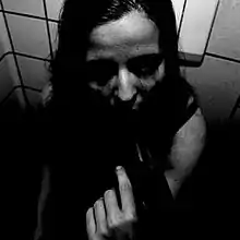 A black and white photograph of a person with their back against a wall who is holding what appears to be gun to their mouth using their left hand. The photograph is in deep shadow and it is difficult to tell if the person is standing or sitting or a man or woman. There appears to be a bra strap on the left shoulder suggesting it is a woman.