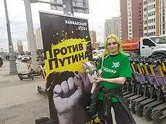 Photographie en couleurs d'une femme portant un T-shirt vert et distribuant des tracts en ville, devant une pancarte politique.