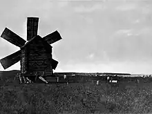 Photographie en noir et blanc d'une plaine avec un moulin en bois à 6 ailes sur la gauche.