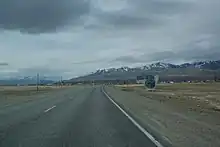 Panneau d'arrivée à Koch-Agatch sur la R256. En arrière-plan, le village se distingue, tandis que des cimes enneigées sont en arrière-plan. Le village se situe dans une steppe plate, celle de la Tchouïa.