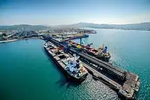 Photographie aérienne d'une jetée d'un port marchand moderne, avec deux cargos à quai de chaque côté de la jetée. La mer est bleu clair, le ciel bleu, et il y a des montagnes en fond.