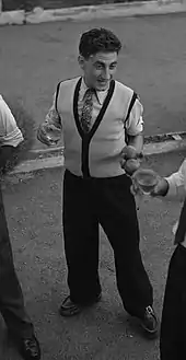 Photographie en noir et blanc d'un homme portant un verre dans sa main droite et deux boules de pétanque dans sa main gauche.