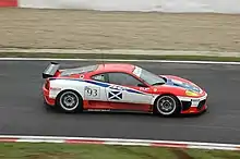 Photographie d'une voiture de grand tourisme blanche, noire, rouge et bleue, vue de haut et de profil, sur une piste.