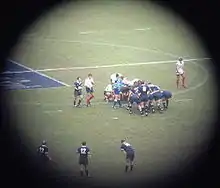 prise de vue lointaine avec un cercle entouré de noir pour se focaliser sur une mêlée du match France - Écosse. Les avants sont en position, les demis en mouvement autour. La phase de jeu n'est pas encore entamée.