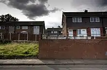 Depuis une rue, vue sur des maisons britanniques du XXe&nbsp;siècle. Les maisons sont surélevées par rapport à la rue. Un terrain vague et herbeux apparaît à la gauche et une mur de briques se trouve à la droite.