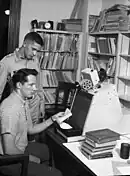 Photographie en noir et blanc de deux personnages concentrés devant un écran, dans une pièce encombrée de documents.