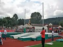 Photo large plan montrant une perchiste en pleine ascension. Des officiels sont au premier plan, la sauteuse se situe au second plan et on aperçoit le public au troisième plan.