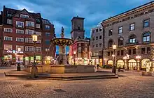 Sur une place bordée de magasins, une fontaine surmontée d'une statue perchée sur un bassin.