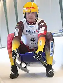 Photo de Felix Loch en bas d'une piste de luge. Il porte une tenue aux couleurs de l'équipe d'Allemagne et par dessus un chasuble blanc. Il porte un casque jaune avec le logo de DHL sur le front, on voit son visage de face à travers la visière de protection. Il soulève l'avant de la luge sur laquelle il est assis.