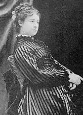 Photographie en noir et blanc d'une femme assise, les cheveux relevés, portant une robe à rayures et des boucles d’oreille