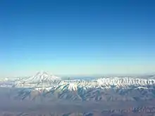 Vue aérienne, crête dominée par un cône volcanique, tous deux enneigés, sous un grand ciel bleu.