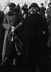Photo en noir et blanc d’un homme en civil, avec cheveux blancs, moustache foncée, lorgnons et tenue sombre, aux côtés d'un militaire à la moustache noire et épaisse ; à l’arrière plan, d’autres civils et militaires