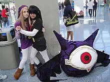 Deux femmes déguisées portant chacune une tenue violette. Au premier plan, on distingue une boule noire et violette ailée, possédant un seul œil géant.