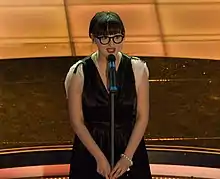 Photographie de face, hauteur trois quarts, d'une jeune femme sur une scène tenant la tige d'un micro sur pied des deux mains, coupe de cheveux « au carré », portant lunettes à bords noirs, lèvres rouges et robe aux bras dénudés, un bracelet à boules blanches sur le poignet gauche.