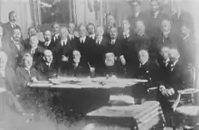 Photographie en noir et blanc d'hommes dans une pièce. Huit sont assis autour d'une table. Une vingtaine plus loin se tiennent debout.