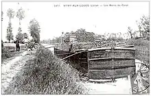 Carte postale noir et blanc du début du siècle. Un bateau, de type bâtard nivernais, une embarcation pouvant transporter 120&nbsp;tonnes de marchandises, est amarré au canal. Il est chargé de rondins de bois. Des ouvriers, à gauche, posent à côté de leur brouette spécifique pour le transport du bois, une brouette n’étant pas fermée sur le côté et pouvant transporter les rondins posés transversalement. Plusieurs planches posées transversalement entre le chemin de halage et le bateau font office de passerelle.