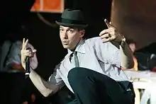 Photo d'un homme blanc sur scène avec une chemise à rayures et un chapeau, qui fait le V de la victoire avec ses deux mains et porte un micro dans la main droite.