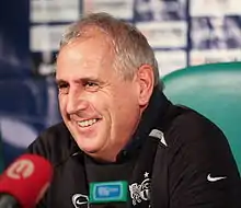 Homme, grisonnant, assis derrière des micros et devant un panneau publicitaire, souriant