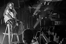 Photo d'une jeune femme assise sur un tabouret installer sur une scène devant une foule de personne.