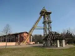 Reconstitution d'une machine à molette datant de 1790 (premier plan), l'ancien chevalement du puits Sainte-Marie (mis en place en février-mars 1988) et la mairie.