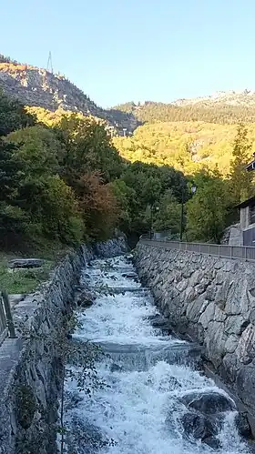 Photographie d'un torrent agité, l'eau étant plus ou moins gelée, contenue dans le canal du torrent en pierres, avec du soleil derrière ainsi que de la forêt.