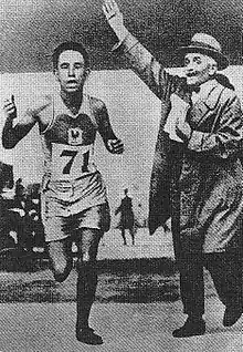 El Ouafi, avec le dossard 71, franchit la ligne d'arrivée devant un juge portant un manteau et coiffé d'un chapeau avec le bas droit levé et des notes dans la main gauche.