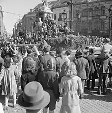 Photo en noir et blanc de la place d'une ville sur laquelle la foule en liesse salue le passage d'un véhicule militaire.