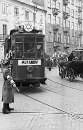 Vu de profil, un homme, coiffé d’un képi et doté d’un brassard, assure la circulation avec un bâton blanc sur une place pavée. Un tramway (sur lequel est peinte une étoile de David et qui porte l’inscription « Muranów » et une automobile circulent devant lui. À l’arrière-plan passent des piétons et cyclistes.