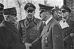 Photo noir et blanc prise le 24 octobre 1940. Au premier plan, Philippe Pétain, chef de l’État français, et Adolf Hitler (tous les deux debout et de profil) échangent une poignée de main. Entre les deux hommes, au second plan, se tient l’interprète officiel de Hitler : Paul-Otto Schmidt. On distingue aussi, au second plan, à droite, le ministre allemand des Affaires étrangères : Joachim von Ribbentrop.