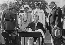 Coolidge assis à une table en extérieur signe un document en présence de plusieurs personnes.
