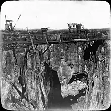 Photographie en noir et blanc d'une falaise d'une carrière à ciel ouvert, avec des échafaudages et des machines-outils sur le bord du trou.