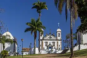 Photographie en couleurs. Vue lointaine de l'église au centre, avec une chapelle à gauche et à droite et des palmiers au premier plan.