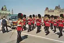 Photo en couleurs d'une fanfare militaire composée de musiciens portant une tunique rouge et un bonnet à poil noir avec une plume jaune jouant d'un instrument à vent défilant sur une rue, un militaire portant une tunique rouge et un bonnet à poil noir avec une plume rouge ayant une épée au fourreau et un bâton à la main marche devant la fanfare ; en arrière-plan, on peut voire des véhicules stationnés, un policier en uniforme et des gens en civil ainsi que des bâtiments derrière