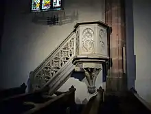 photographie en couleur montrant une petite tribune octogonale en grès gris adossée à un pilier en grès rose et accessible par une escalier doté d’une balustrade en pierre décorée de motifs géométriques