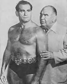 Photo en noir et blanc de deux hommes. L'un est devant l'autre, c'est Lou Thesz, en slip de catch noir et portant une ceinture de champion, et regardant au loin, vers la droite. Il a les cheveux coiffés en arrière. A droite, derrière lui, se tien Ed « Strangler » Lewis en costume clair avec une chemise et pas de cravate avec une main sur le bras gauche du premier et lui disant quelque chose à l'oreille. Il a des cheveux grisonnants rabattus vers l'arrière.