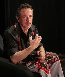 La photo montre Clive Barker  parlant avec animation pendant une conférence. Il porte une chemise et un pantalon sombres et des bretelles rouges.