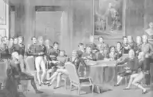 Gravure en nuances de gris, présentant une assemblée d'hommes, en uniformes couverts de décorations, dans une large pièce décorée d'un tableau et d'un buste. Certains, au centre et à droite, sont assis autour d'une table ronde ornée d'une nappe et sur laquelle plusieurs papiers sont posés. Parmi ceux assis au premier plan, cinq personnages : deux à droite, deux au centre, un à gauche. Deux autres sont assis au second plan, derrière la table. De nombreux autres sont debout, à gauche et au second plan.