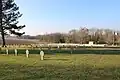 Vue générale sur le cimetière herbeux avec ses nombreuses stèles et croix alignées. Un drapeau français flotte sur un mat. Dans le fond, des arbres et un grand champ labouré.