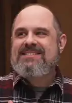 Un homme de 51 ans avec une barbe grise souriant à la gauche de la caméra.