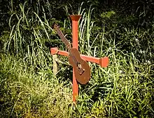 Photographie en couleur d'une croix orange sur lequel est fixé une guitare stylisée dans un matériau qui ressemble à du contreplaqué. La croix est plantée dans un sol recouvert de végétation.
