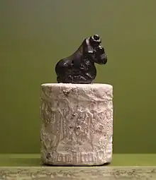 Cylindre gravé de couleur claire, surmonté par une figurine d'un bélier accroupi.