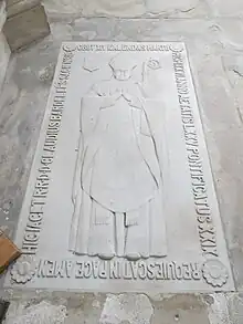 Photo de longue dalle sculptée en bas relief représentant un évêque mitré, avec inscription funéraire sur le pourtour.