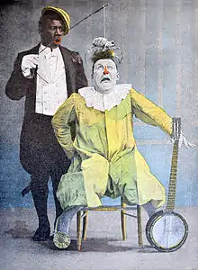 Illustration en couleurs montrant un clown noir posant une araignée au bout d'une baguette sur la tête d'un autre clown vêtu de jaune assis devant lui et tenant un banjo dans sa main gauche.